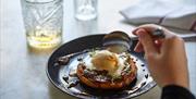 A black plate with eggs benedict on it - a hand is reaching into the food holding a spoon