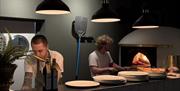 A kitchen with 2 chefs behind it, and there are pizza plates atop of the counter