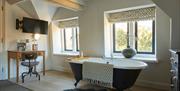 A free standing bathtub with a rug hanging over the side. There is a desk and a tv at the end of the bath