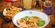 A bowl of chicken ramen with an egg, alongside a plate of gyozas, jar of chilli oil and a drink.