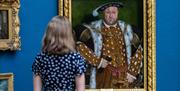 A lady looking at a painting of King Henry VIII in a gold frame on a blue wall.