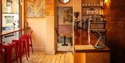A side view of an empty wooden bar to the right, and a row of empty bar stools facing a wall-length window to the right.