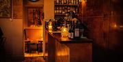 A side view of a bar containing several bottles of wine with warm lighting. Two shelves containing alcohol bottles are visible behind.