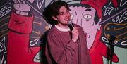 A comedian with a microphone in a burgundy sweatshirt, on stage against a Jester cartoon backdrop