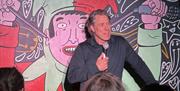A comedian with a microphone in a black shirt, on stage against a Jester cartoon backdrop