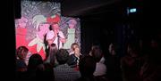 A comedian with a microphone in a white t-shirt, on stage against a Jester cartoon backdrop and people in the crowd