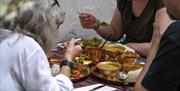A group of people eating a selection of Nepalese dishes around a table with a brightly coloured striped tablecloth, in an outdoor dining setting.