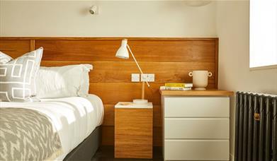 A white bed, gray and white pillows, a wooden headboard, white and wooden nightstands, a white lamp, a beige vase, yellow books, and sunlight streamin