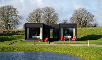 Modern-style black and white accommodation building with red chairs in front, overlooking a lake.
