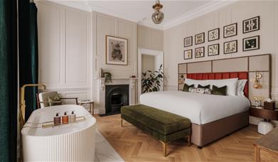 Highly spec'ed bedroom with oak parquet flooring, a green velvet ottoman and free standing bath at the end of a double bed with a Victorian fireplace.