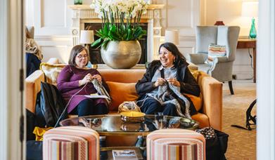 Two women knitting while sitting in a lounge on armchairs.
